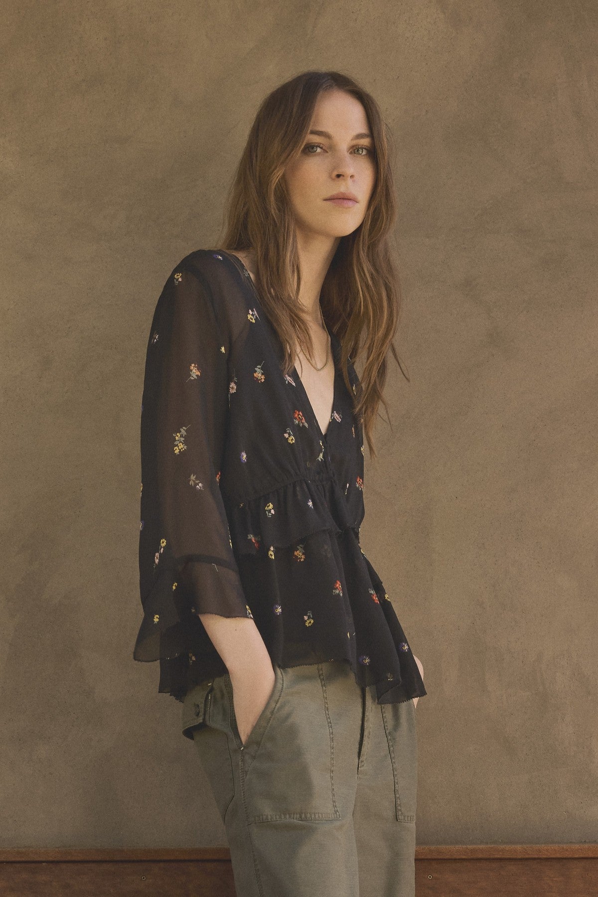 A woman with long brown hair wears the AILEEN BLOUSE by Velvet by Graham & Spencer—a sheer viscose floral print top—paired with olive green pants, standing against a textured beige wall.