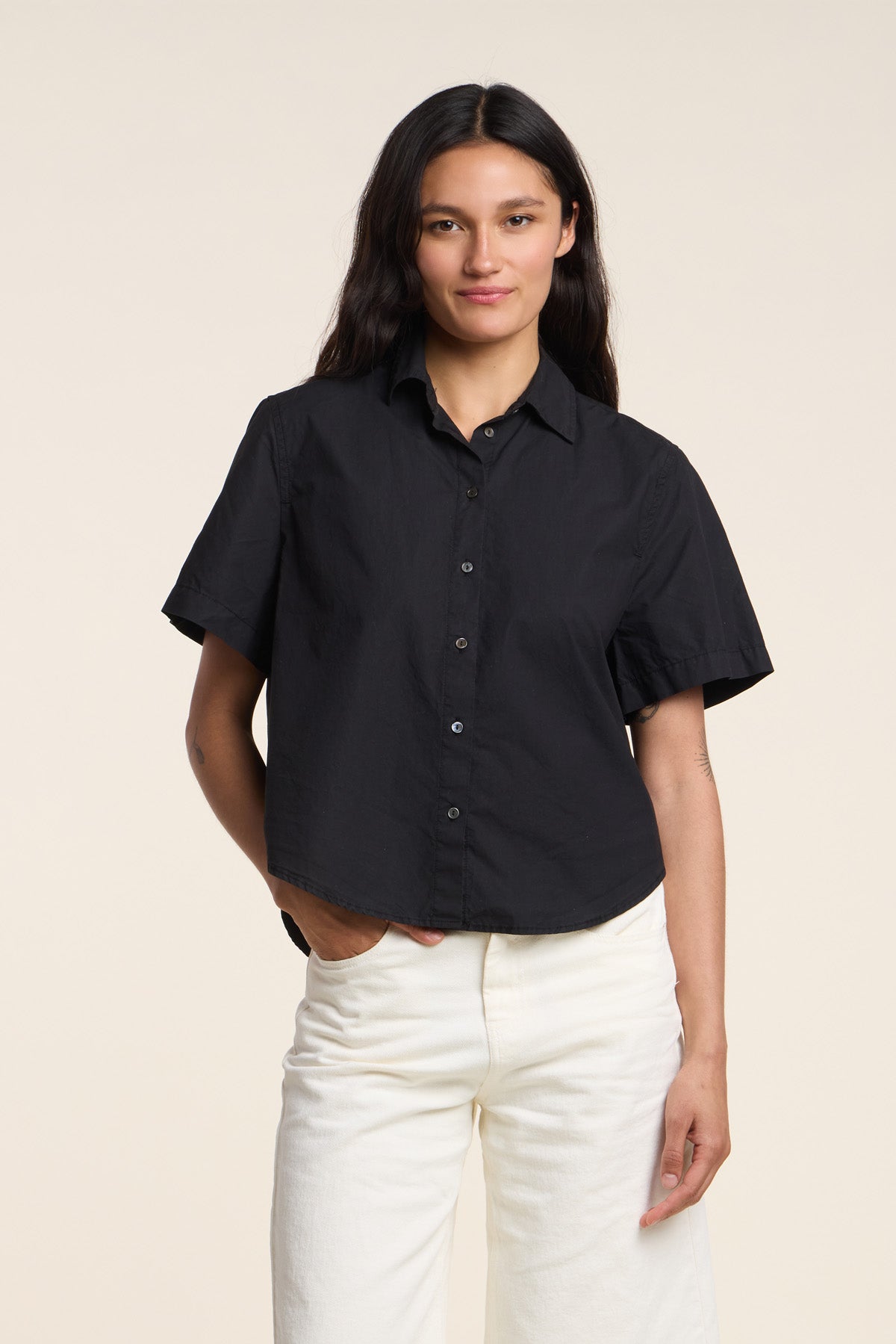 A person stands against a plain light background wearing the ALANA SHIRT by Velvet by Graham & Spencer— a short-sleeved black cotton poplin shirt with a classic collar—paired with white pants.