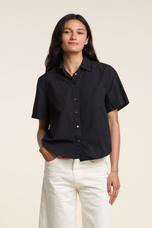 A person wearing the ALANA SHIRT by Velvet by Graham & Spencer—a women’s short sleeve black cotton poplin shirt—and white pants stands against a plain light background with one hand in their pocket.