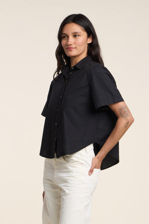 Model wearing the ALANA SHIRT by Velvet by Graham & Spencer—a black cotton poplin shirt with a classic collar and short sleeves—paired with white pants, hands in pockets, posed against a plain background.
