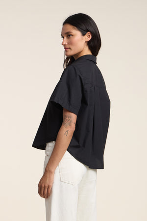 A woman in profile wears the ALANA SHIRT by Velvet by Graham & Spencer—a loose cotton poplin short sleeve shirt—paired with white pants, showing a tattoo on her left arm against a plain background.