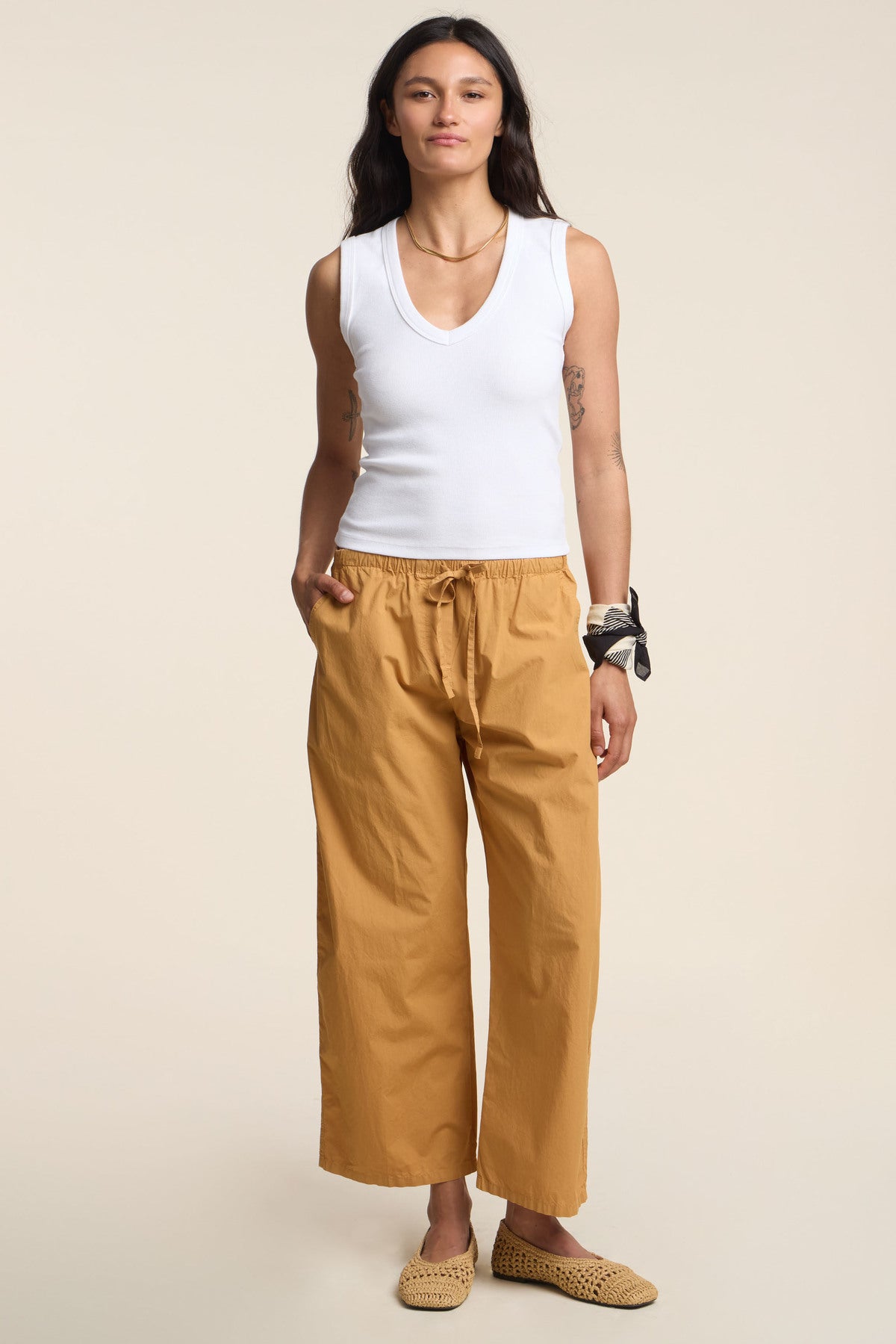 A person stands against a plain background wearing a white sleeveless top, Velvet by Graham & Spencer's ALLEGRA PANT in cotton poplin with a cropped barrel leg, woven slip-on shoes, and a bandana tied around their wrist.-41384062222529