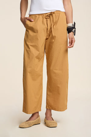 Person wearing the ALLEGRA PANT by Velvet by Graham & Spencer in mustard yellow cotton poplin, styled with a white top, straw slip-ons, and a black-and-white bandana on their wrist.