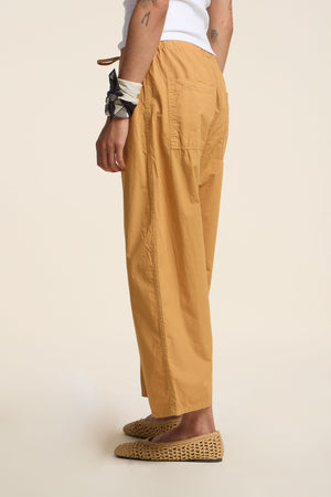 Wearing Velvet by Graham & Spencer’s ALLEGRA PANT in mustard yellow cotton poplin with a wide barrel leg and cropped fit, styled with a white sleeveless top, woven slip-ons, and a black-and-white bandana on the left wrist; shown in profile.