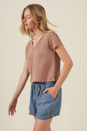 A woman wearing the AMBER TEE by Velvet by Graham & Spencer—a ribbed V-neck brown linen knit top—paired with blue drawstring shorts stands against a plain light background.