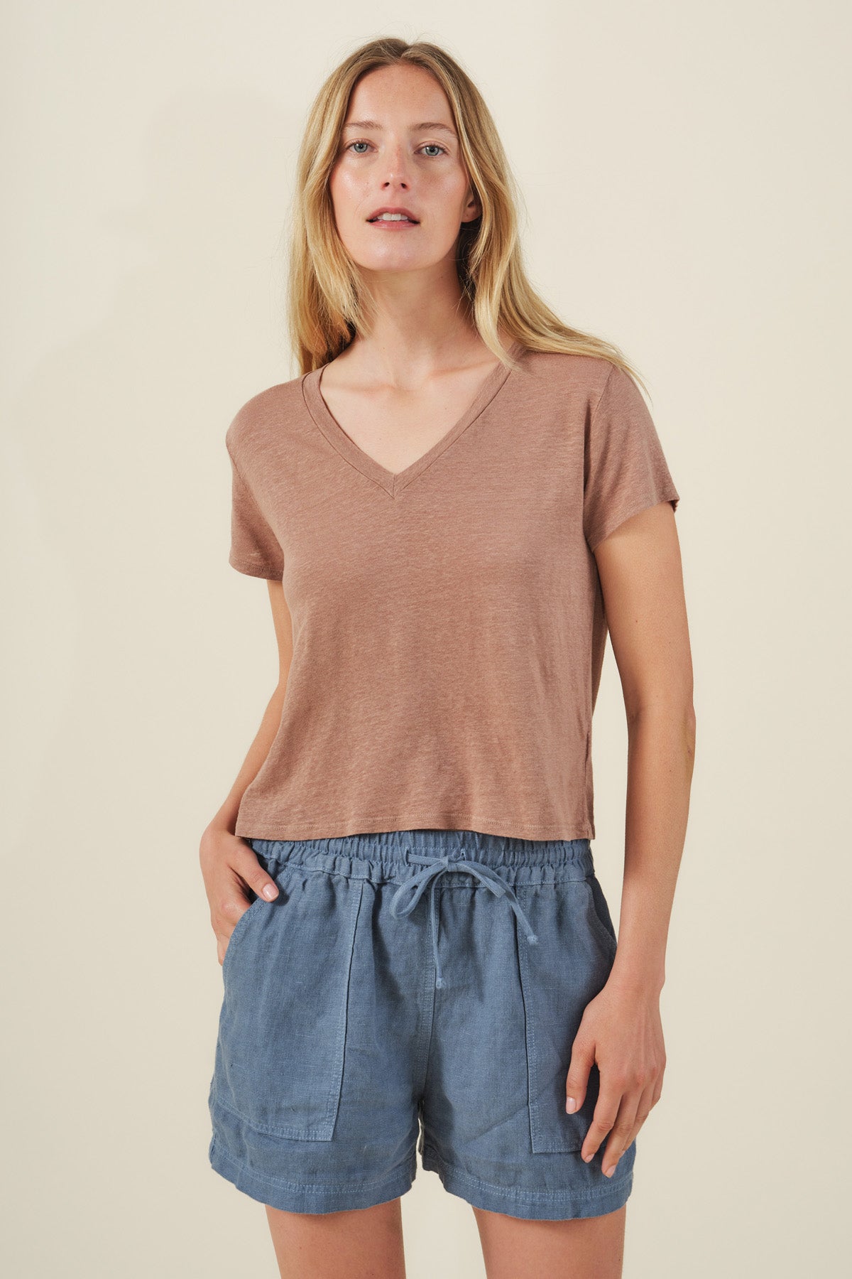 A woman stands against a plain background wearing the AMBER TEE by Velvet by Graham & Spencer—a light brown linen knit top with a ribbed V-neckline—paired with blue drawstring shorts, her hands in her pockets.