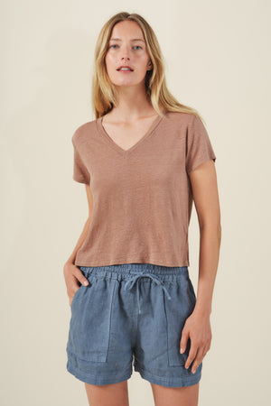 A woman stands against a plain background wearing the AMBER TEE by Velvet by Graham & Spencer—a light brown linen knit top with a ribbed V-neckline—paired with blue drawstring shorts, her hands in her pockets.