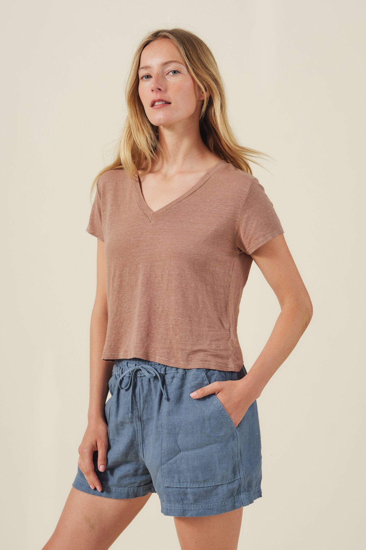 A woman poses with one hand in her pocket, wearing the AMBER TEE by Velvet by Graham & Spencer—a light brown ribbed V-neckline linen knit—paired with blue drawstring shorts against a plain background.-41526765060289