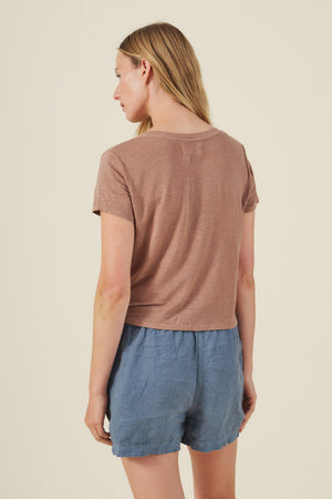 A person with long blonde hair is seen from behind, wearing the Velvet by Graham & Spencer AMBER TEE—a brown linen knit shirt with a ribbed V-neckline—paired with blue shorts against a plain background.