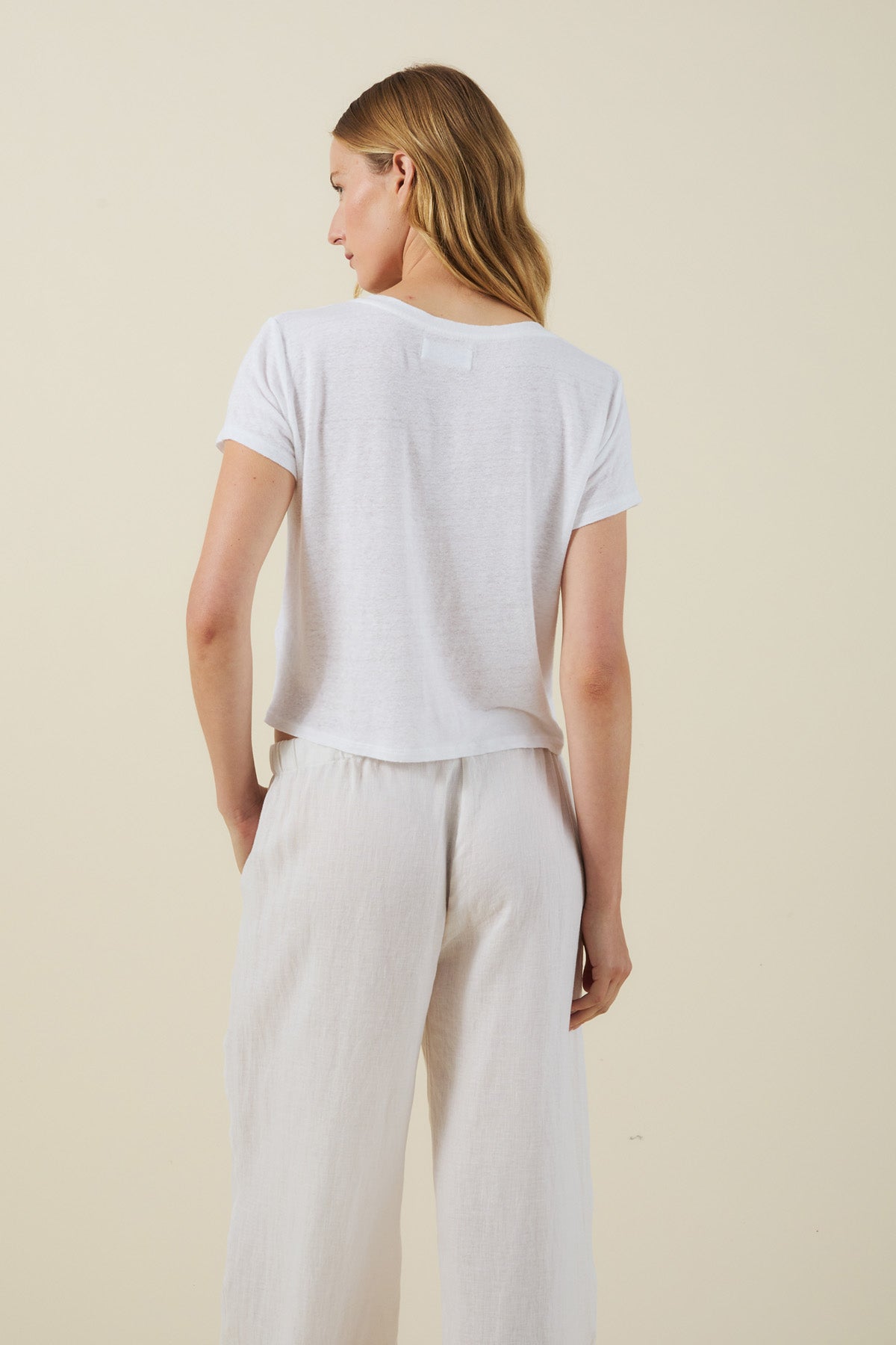 A person stands facing away, wearing the AMBER TEE by Velvet by Graham & Spencer—a white cropped boxy fit shirt—paired with loose white pants against a plain light background.