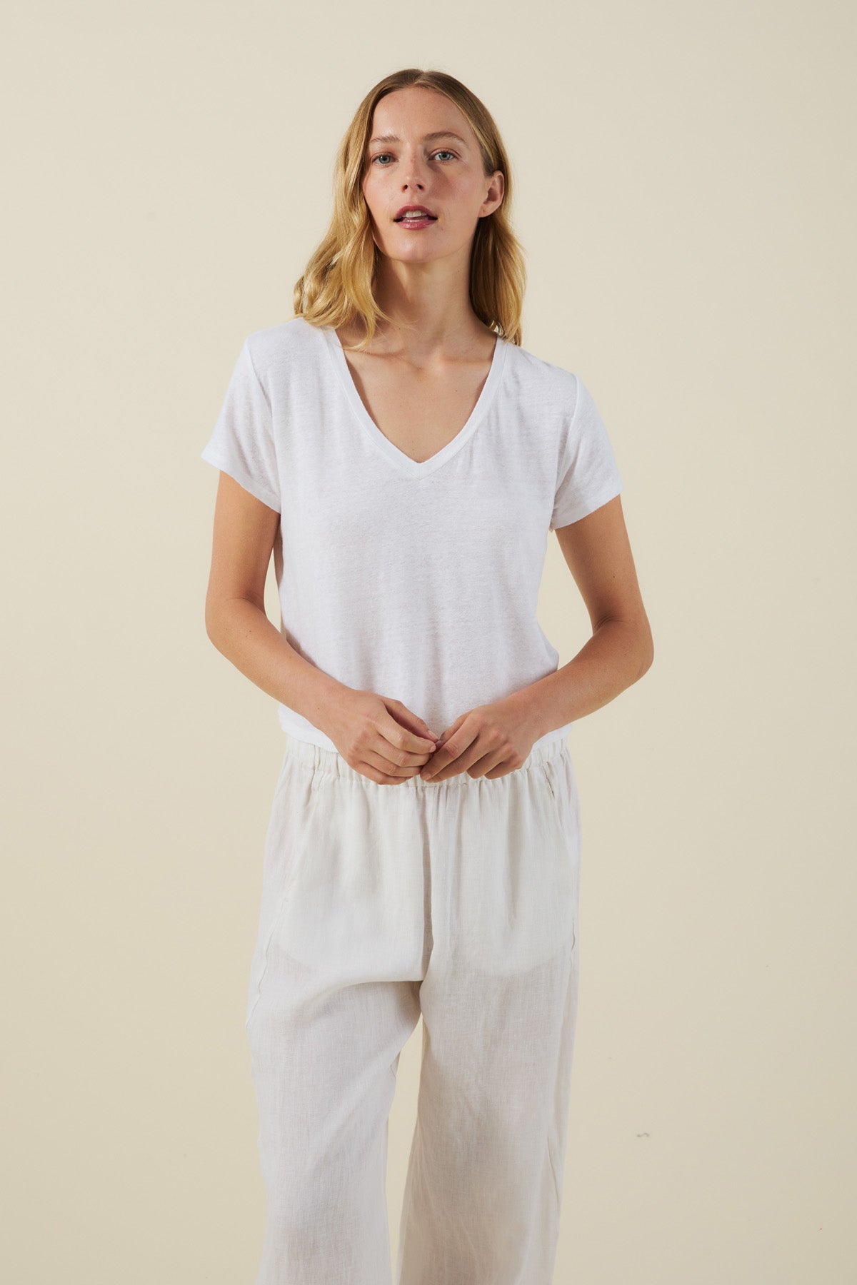 A woman stands against a plain background wearing the AMBER TEE by Velvet by Graham & Spencer, a white linen knit blend V-neck T-shirt, paired with loose white pants and her hands clasped in front of her.