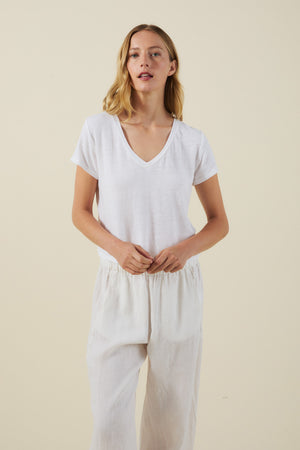 A woman stands against a plain background wearing the AMBER TEE by Velvet by Graham & Spencer, a white linen knit blend V-neck T-shirt, paired with loose white pants and her hands clasped in front of her.
