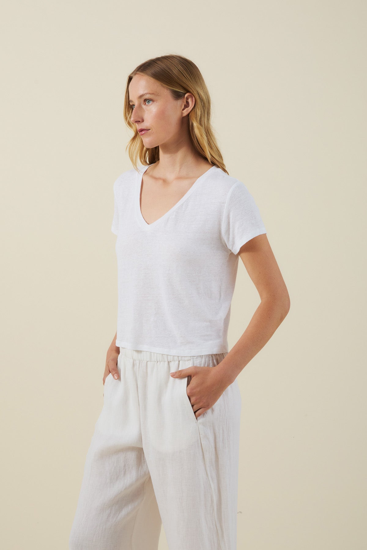 A woman in the AMBER TEE by Velvet by Graham & Spencer, a white linen knit blend V-neck T-shirt, and white drawstring pants stands with her hands in her pockets against a plain light background.-41502226710721