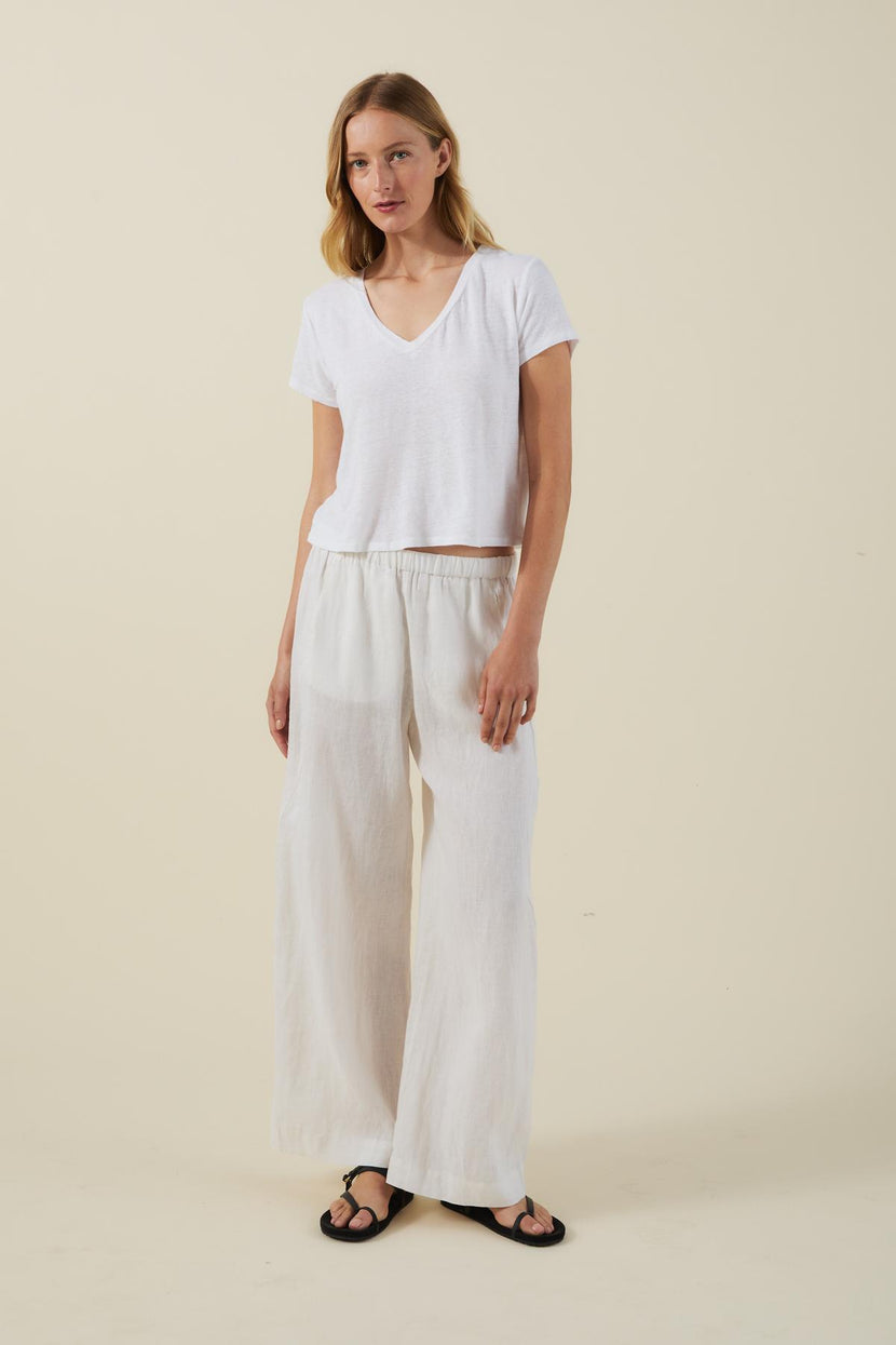 A woman stands against a plain background wearing the AMBER TEE by Velvet by Graham & Spencer, paired with loose white pants and black sandals.