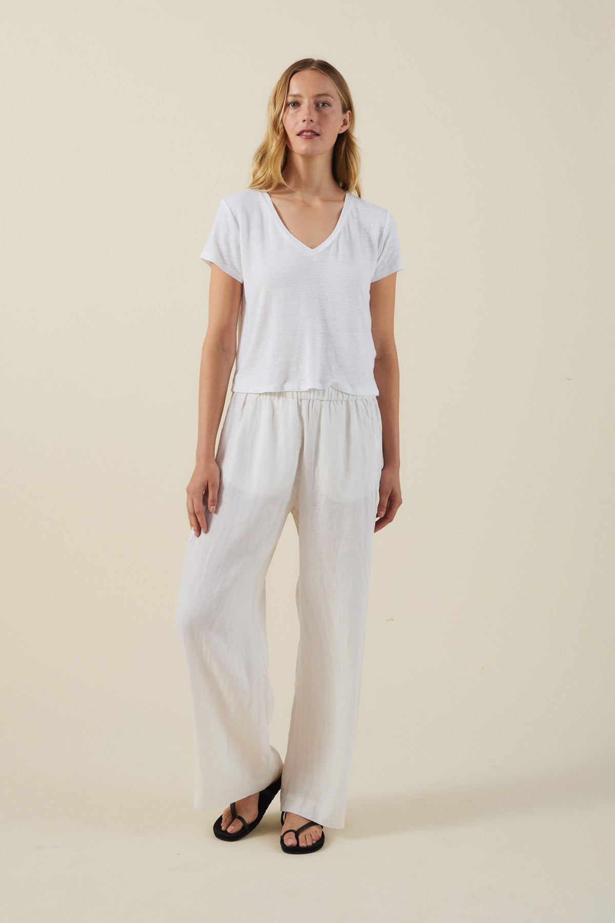 A woman stands against a plain background wearing a white V-neck T-shirt, black sandals, and the STEPHANIE LINEN PANT by Velvet by Graham & Spencer—wide-leg linen pants with an elastic waist.