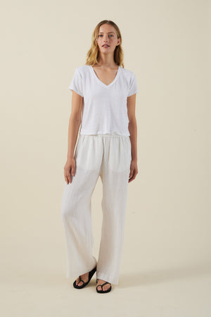 A woman stands against a plain background wearing a white V-neck T-shirt, black sandals, and the STEPHANIE LINEN PANT by Velvet by Graham & Spencer—wide-leg linen pants with an elastic waist.