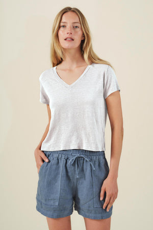 A woman stands against a plain background wearing the AMBER TEE by Velvet by Graham & Spencer, a light gray ribbed V-neck T-shirt, paired with blue drawstring shorts and her hands in her pockets.