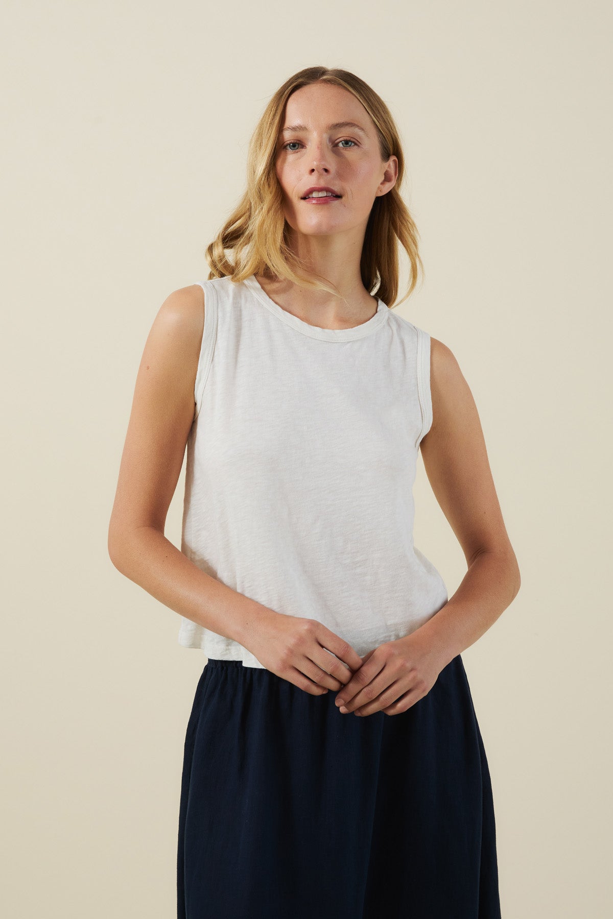 A woman stands against a plain background, wearing the white AMBRIA TANK TOP by Velvet by Graham & Spencer and a dark skirt, her hands resting gently in front of her.