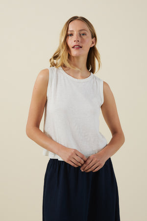 A woman stands against a plain background, wearing the white AMBRIA TANK TOP by Velvet by Graham & Spencer and a dark skirt, her hands resting gently in front of her.