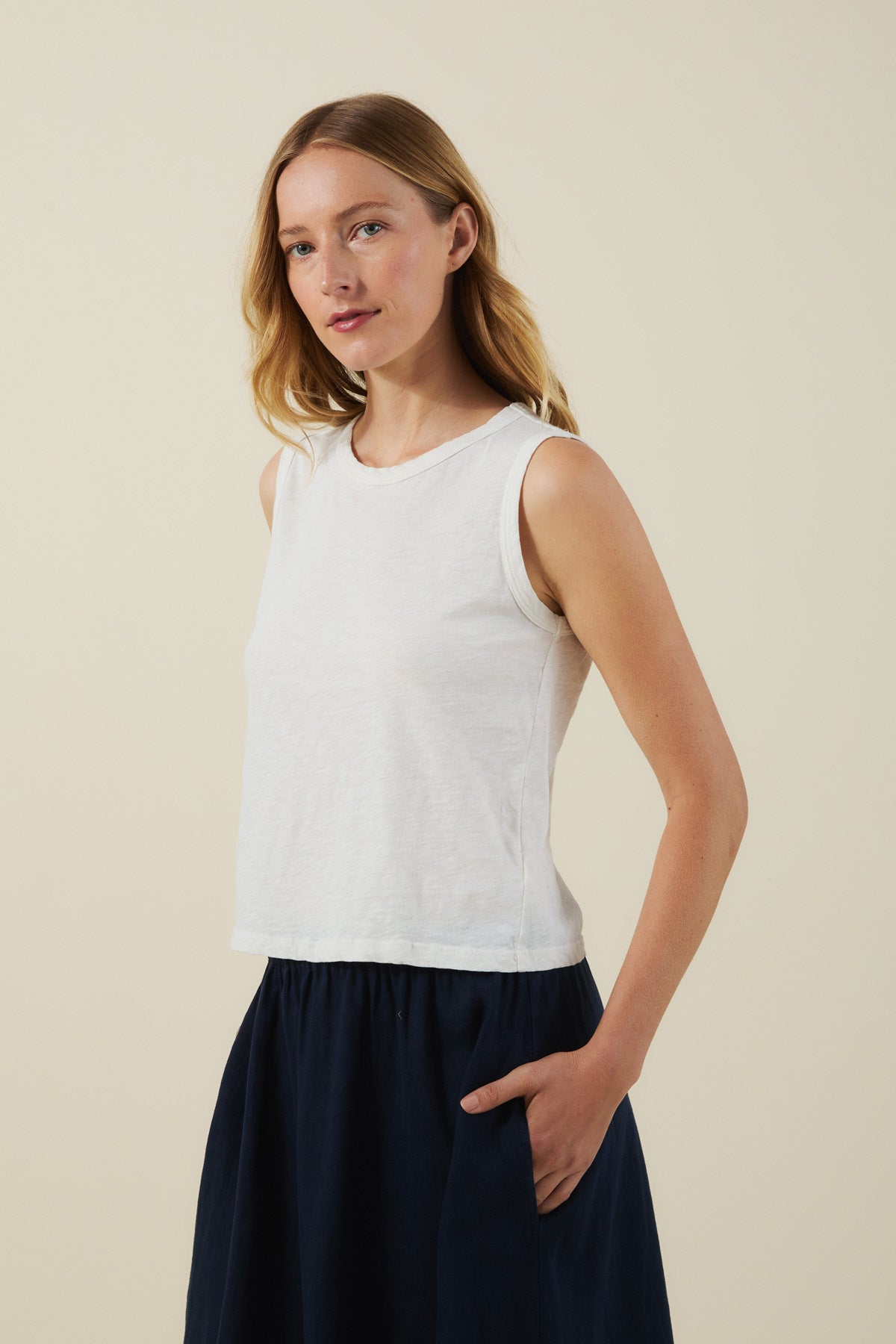 A woman wears the sleeveless white AMBRIA TANK TOP by Velvet by Graham & Spencer with a dark skirt, standing against a plain light background, one hand in her pocket.-41454618935489
