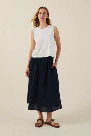 A woman stands against a plain background wearing the AMBRIA TANK TOP by Velvet by Graham & Spencer—a white textured cotton slub sleeveless style—paired with a navy midi skirt and brown sandals, creating an effortless summer look.