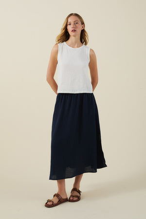 A woman stands against a plain background wearing a white sleeveless top, brown sandals, and the VIRGINIA LINEN SKIRT by Velvet by Graham & Spencer—an A-line midi skirt with an elastic waist.