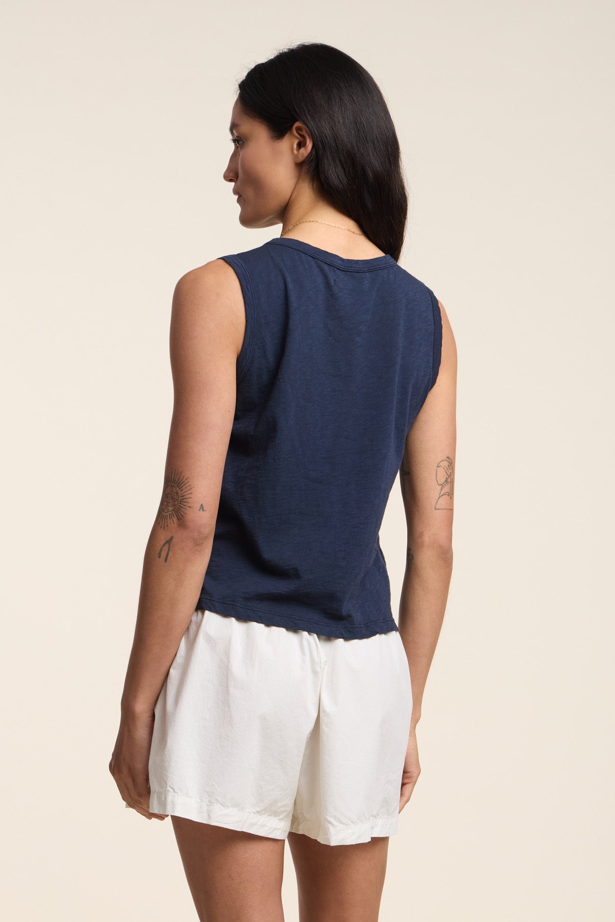 A person with long dark hair is shown from the back, revealing arm tattoos, wearing Velvet by Graham & Spencer's AMBRIA TANK TOP in navy—a boxy, cropped summer style—paired with white shorts.