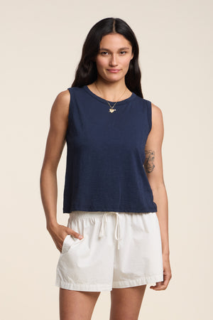 A person stands facing forward, wearing the AMBRIA TANK TOP by Velvet by Graham & Spencer in navy cotton slub and white drawstring shorts, with one hand in a pocket, against a plain background—a laid-back ensemble ideal for your summer wardrobe.