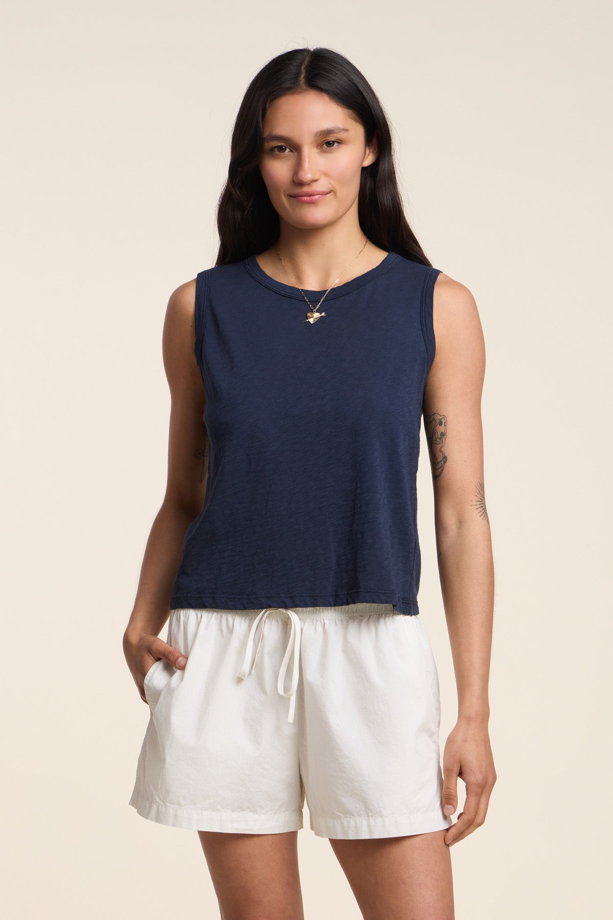 A woman stands against a plain background wearing the Velvet by Graham & Spencer AMBRIA TANK TOP, a navy cropped summer top with a boxy swing silhouette, paired with white drawstring shorts and one hand in her pocket.