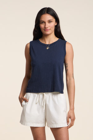 A woman stands against a plain background wearing the Velvet by Graham & Spencer AMBRIA TANK TOP, a navy cropped summer top with a boxy swing silhouette, paired with white drawstring shorts and one hand in her pocket.