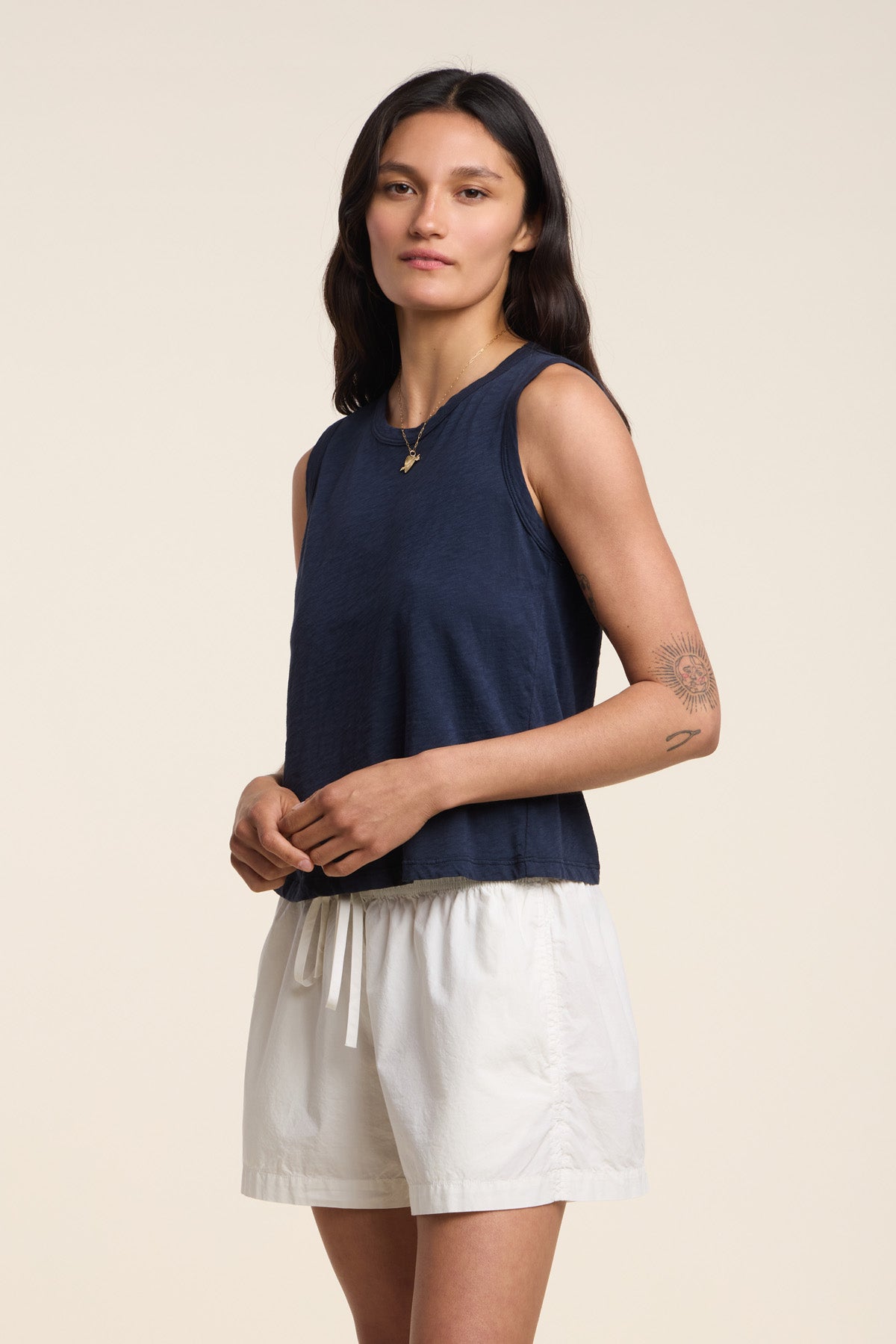 Someone stands against a plain light background wearing the AMBRIA TANK TOP by Velvet by Graham & Spencer—a sleeveless, boxy swing silhouette navy cotton slub top—paired with white shorts.
