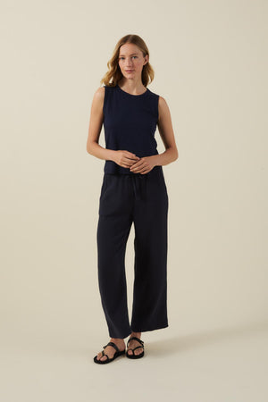 A woman stands against a plain background wearing a navy sleeveless top, Velvet by Graham & Spencer's FRANNY PANT in cotton gauze with a pull-on waistband, and black sandals.