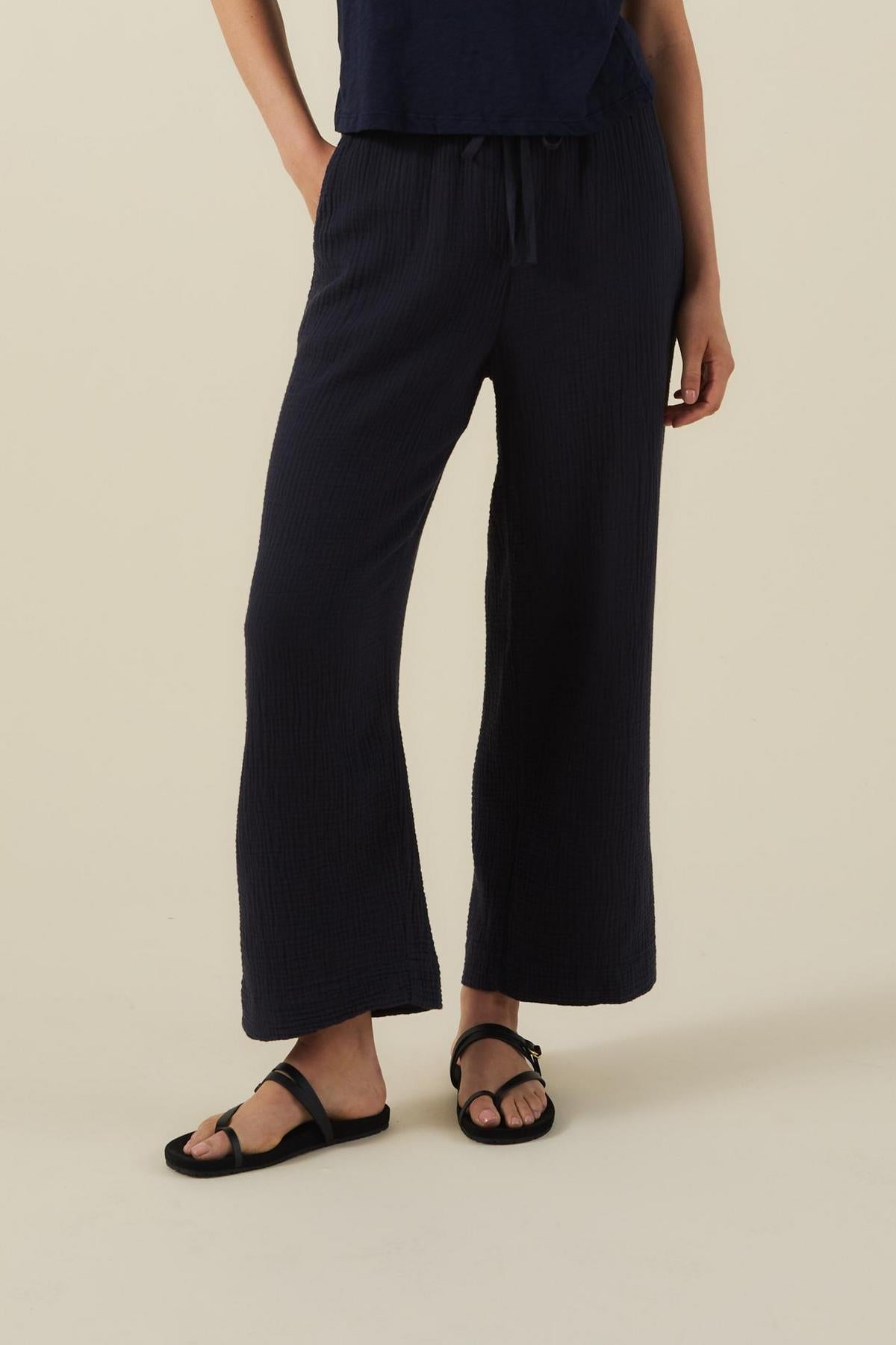 A person wears the FRANNY PANT by Velvet by Graham & Spencer—loose-fitting, navy blue cotton gauze pants with a pull-on waistband—paired with black sandals and a navy top, standing against a beige background.-41527347118273