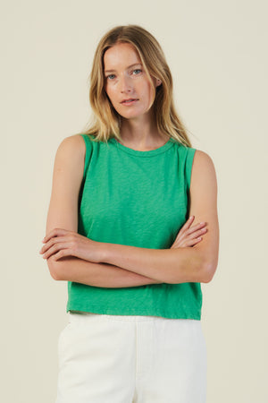 A woman with long blonde hair models the AMBRIA TANK TOP by Velvet by Graham & Spencer, a cropped summer style with a boxy swing silhouette, paired with white pants and posed against a plain background.