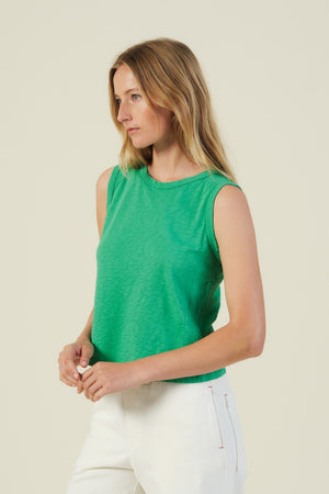 A woman models the AMBRIA TANK TOP by Velvet by Graham & Spencer—a sleeveless green boxy swing top—paired with white pants, standing against a plain light background and looking to the side.