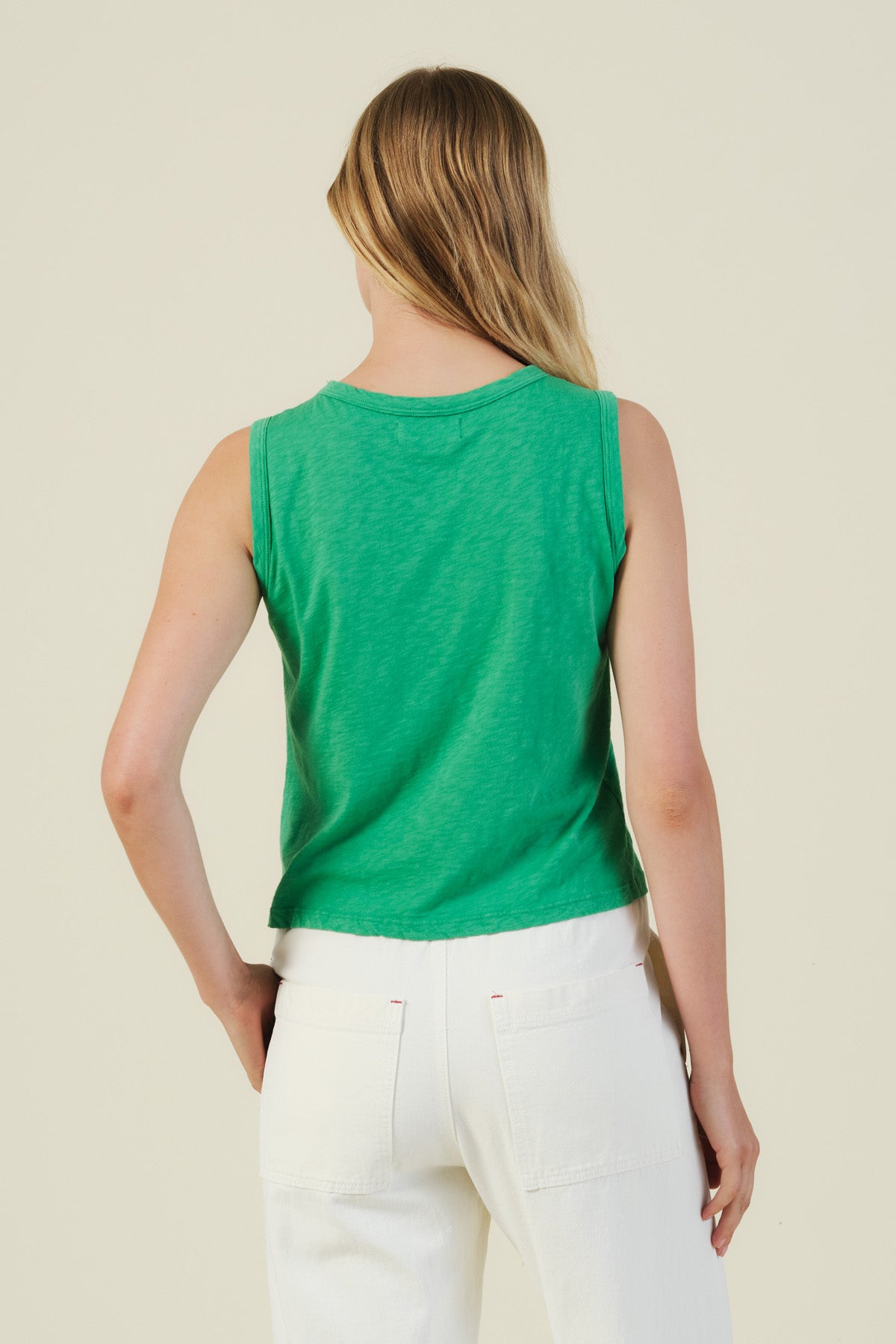 A person with long, light brown hair is seen from the back wearing the AMBRIA TANK TOP by Velvet by Graham & Spencer—a boxy, sleeveless green top—paired with high-waisted white pants against a plain light background.