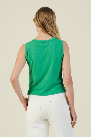 A person with long, light brown hair is seen from the back wearing the AMBRIA TANK TOP by Velvet by Graham & Spencer—a boxy, sleeveless green top—paired with high-waisted white pants against a plain light background.