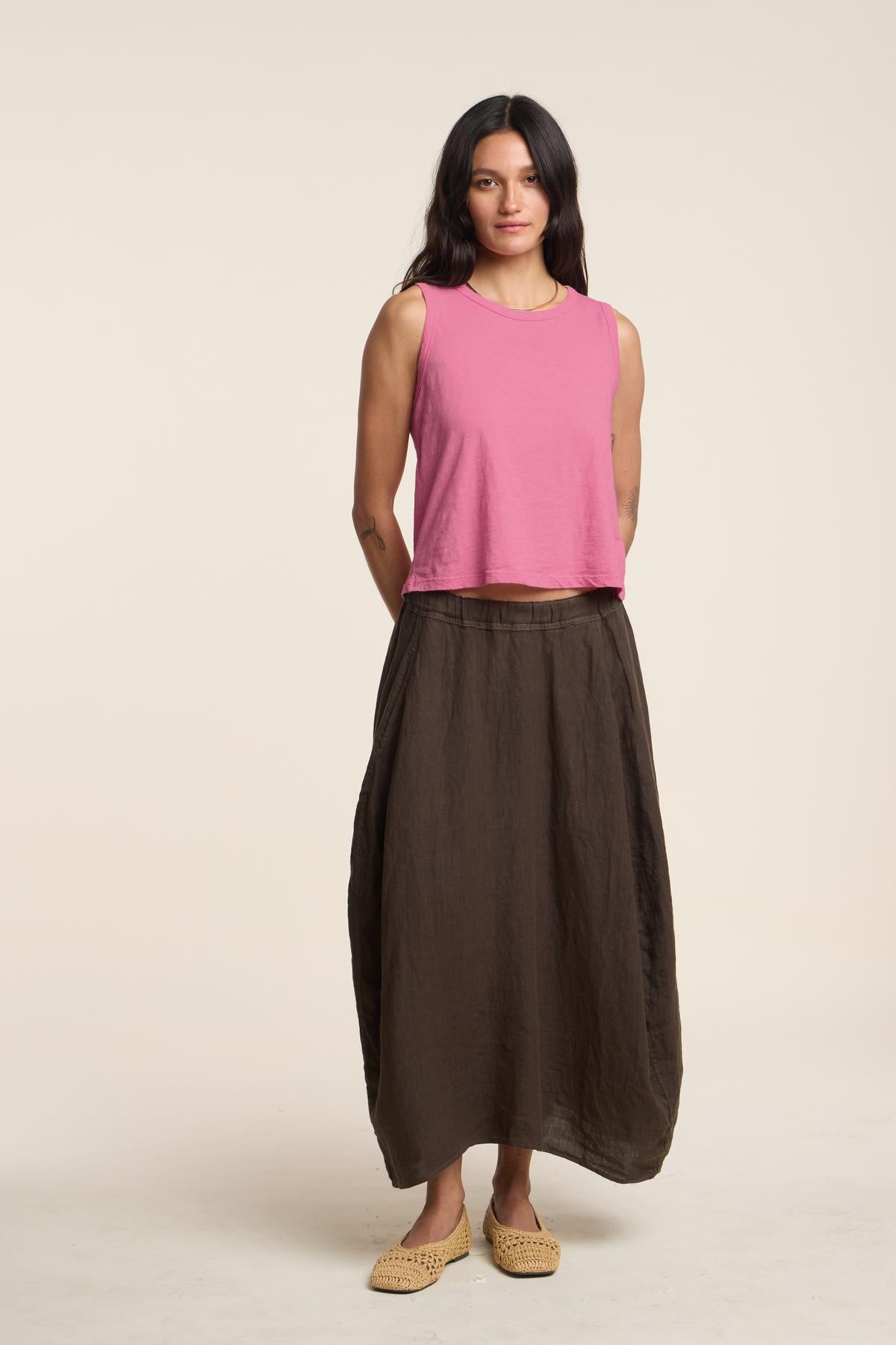 A woman faces forward against a plain light background, wearing the FAE LINEN SKIRT by Velvet by Graham & Spencer—a long brown A-line linen skirt—paired with a pink sleeveless top and beige woven shoes.-41460565868737