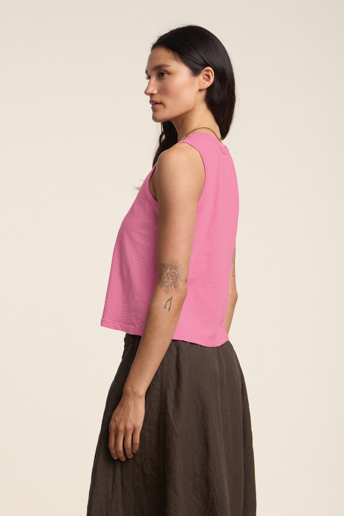 A woman with long dark hair and visible upper arm tattoos stands sideways against a plain light background, wearing the AMBRIA TANK TOP in pink by Velvet by Graham & Spencer and a brown skirt.-41387989270721