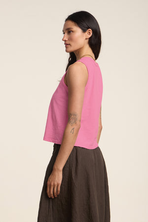 A woman with long dark hair and visible upper arm tattoos stands sideways against a plain light background, wearing the AMBRIA TANK TOP in pink by Velvet by Graham & Spencer and a brown skirt.