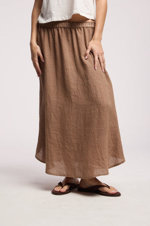A person stands against a plain light background wearing a white short-sleeve top, dark flip-flops, and the VIRGINIA LINEN SKIRT by Velvet by Graham & Spencer—a loose, woven linen A-line midi skirt with an elastic waist.