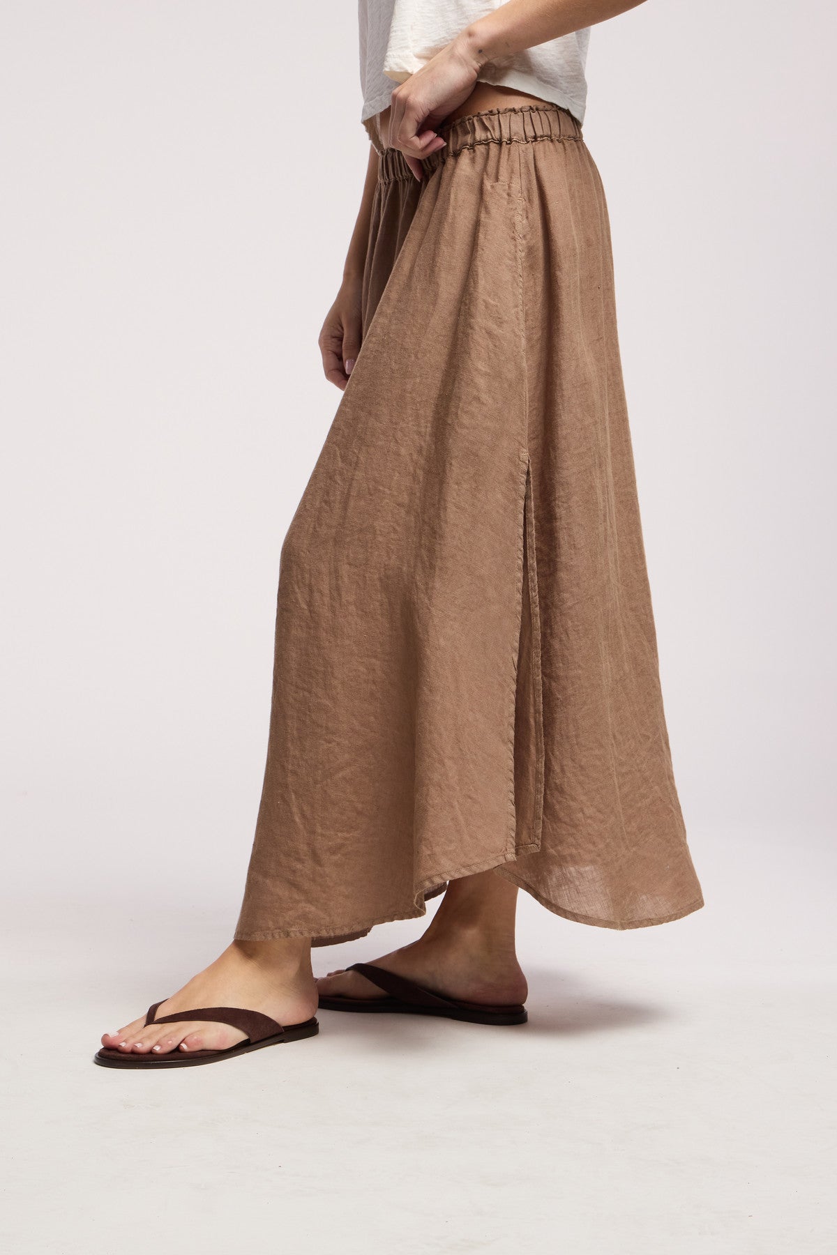 A person wears the VIRGINIA LINEN SKIRT by Velvet by Graham & Spencer, a light brown woven linen skirt with an elastic waistband and subtle slit, styled with a white top and brown flip-flops.
