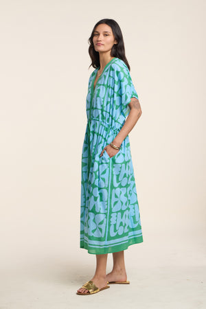 A woman stands sideways wearing the AMIRA DRESS by Velvet by Graham & Spencer—a loose-fitting, blue and green patterned cotton kaftan with short sleeves—paired with sandals against a plain light background.