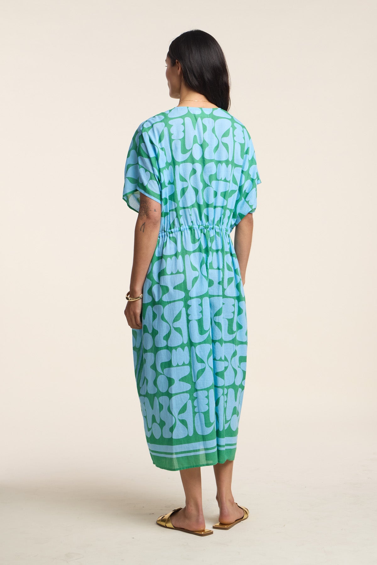 A woman stands with her back to the camera, wearing the AMIRA DRESS by Velvet by Graham & Spencer—a loose blue and green patterned cotton kaftan—paired with flat sandals for an effortlessly travel-ready look.