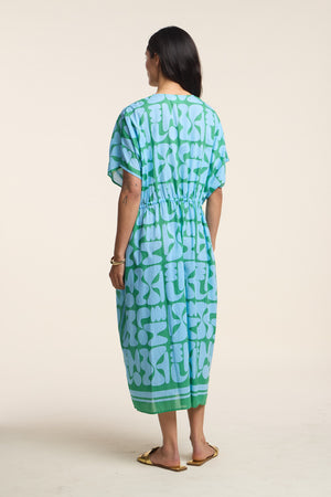 A woman stands with her back to the camera, wearing the AMIRA DRESS by Velvet by Graham & Spencer—a loose blue and green patterned cotton kaftan—paired with flat sandals for an effortlessly travel-ready look.