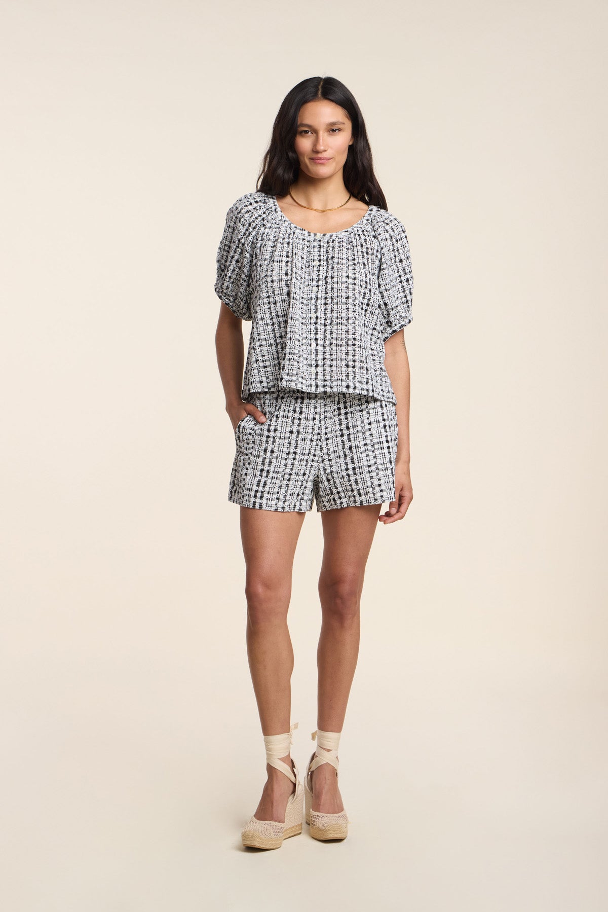 A woman stands wearing the DANIELLA SHORT set by Velvet by Graham & Spencer, featuring a matching short-sleeve blouse and blue and white gingham shorts, paired with light espadrille wedges.-41526918774977