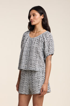 A woman stands in profile wearing the ANGIE TOP by Velvet by Graham & Spencer paired with matching shorts, a simple necklace, and loose hair against a plain background.