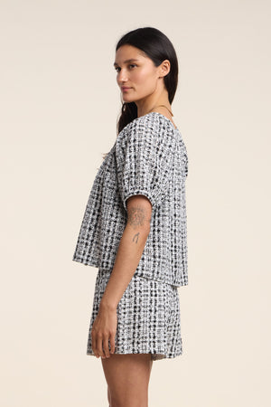 A woman in profile wears the Velvet by Graham & Spencer ANGIE TOP in black and white gingham, paired with matching shorts and displays a visible upper arm tattoo against a plain background.