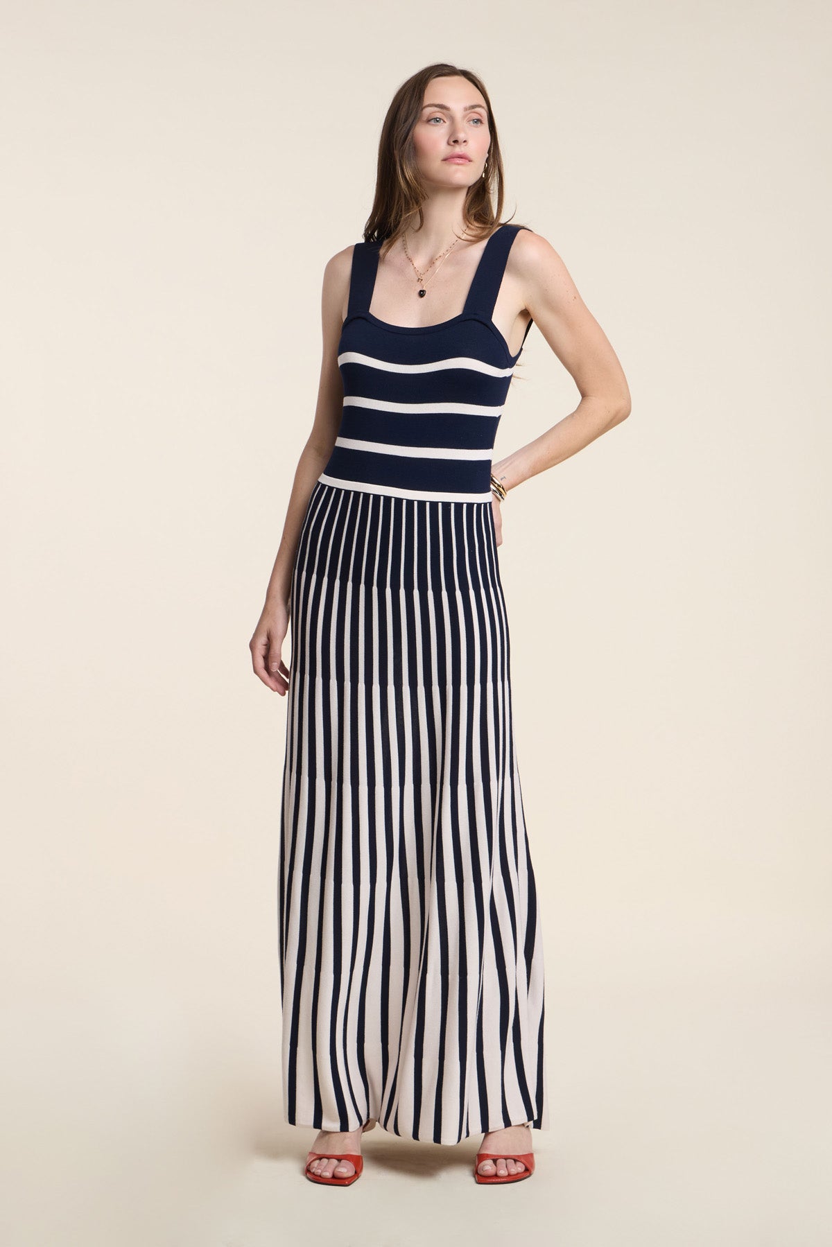 A woman wears the AVELINE DRESS by Velvet by Graham & Spencer—a sleeveless navy and white striped maxi with a fitted bodice and pleated skirt—paired with red sandals and a necklace, set against a beige background.-41526920446145