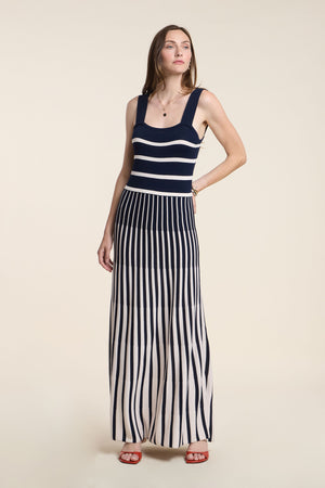 A woman wears the AVELINE DRESS by Velvet by Graham & Spencer—a sleeveless navy and white striped maxi with a fitted bodice and pleated skirt—paired with red sandals and a necklace, set against a beige background.